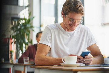 Smiling man with headphones using cell phone and drinking coffee in cafe