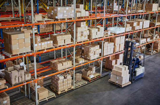 Worker operating forklift moving cardboard boxes along distribution warehouse shelves