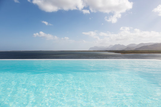 Sunny, tranquil infinity pool with ocean view under blue sky with clouds
