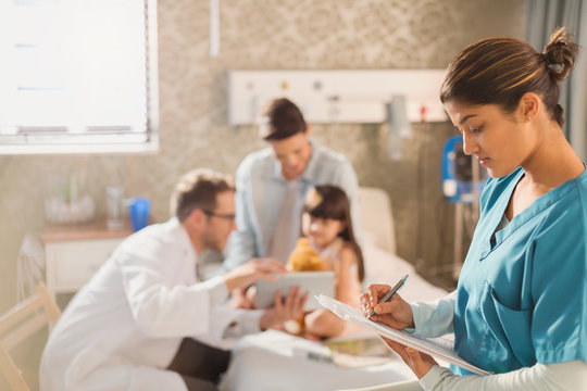Female nurse taking notes on medical record clipboard while doctor shows digital tablet to girl patient in hospital