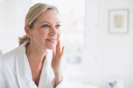 Smiling mature woman applying moisturizer to face at bathroom mirror