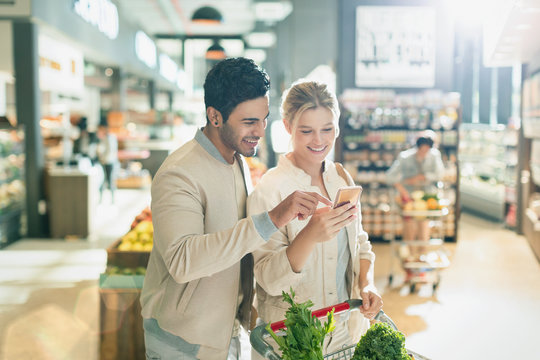 Young couple using cell phone, grocery shopping in grocery store market - Powered by Adobe