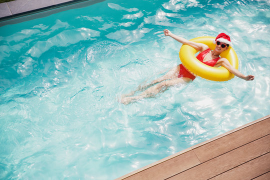Portrait carefree woman in Santa hat and heart-shape sunglasses swimming with inflatable ring in swimming pool