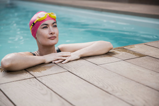Confident woman in swimming cap and goggles leaning on edge of swimming pool