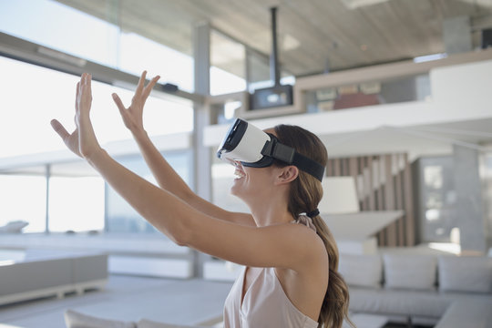 Smiling woman using virtual reality simulator glasses with arms raised in modern, luxury home showcase living room
