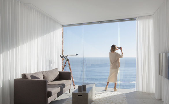 Woman with camera phone photographing sunny ocean view from modern, luxury home showcase interior living room