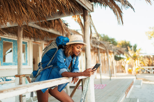Young female backpacker using smart phone on beach hut patio