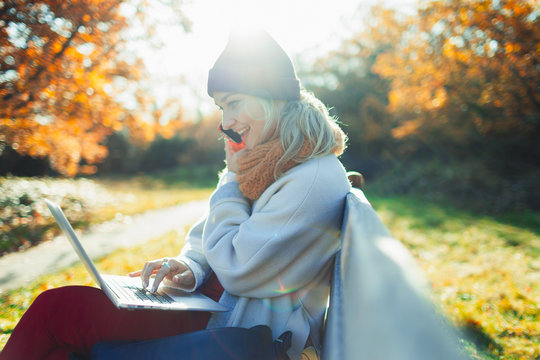 Young woman using laptop and talking on smart phone on sunny autumn park bench