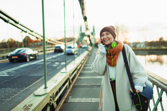 Young woman in stocking cap and scarf talking on smart phone on bridge