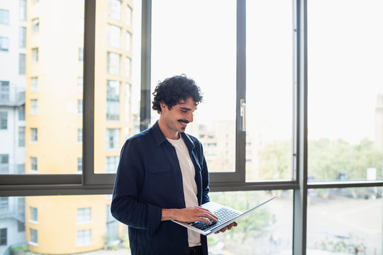 Smiling man using laptop at urban apartment window