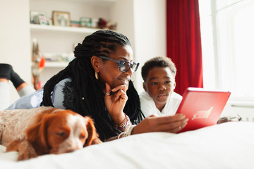 Grandmother and grandson using digital tablet next to sleeping dog on bed