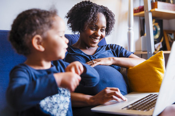 Happy pregnant woman with daughter using laptop on sofa