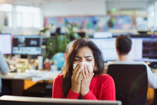 Businesswoman with allergies sneezing into tissue at computer in office