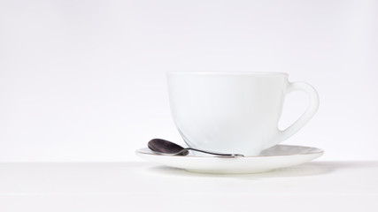 A white Cup for tea or coffee and a metal spoon on a white table on a gray background.