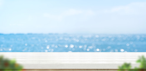 Empty white plank wood table with blur blue sky and sea bokeh background with green leaf foreground,Mock up template for display or montage of product or content use as banner in social media ads