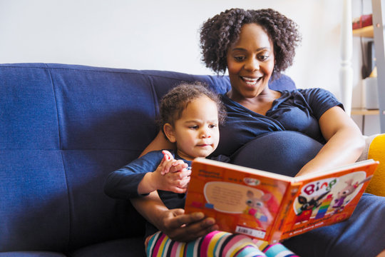 Pregnant mother reading book to daughter on sofa