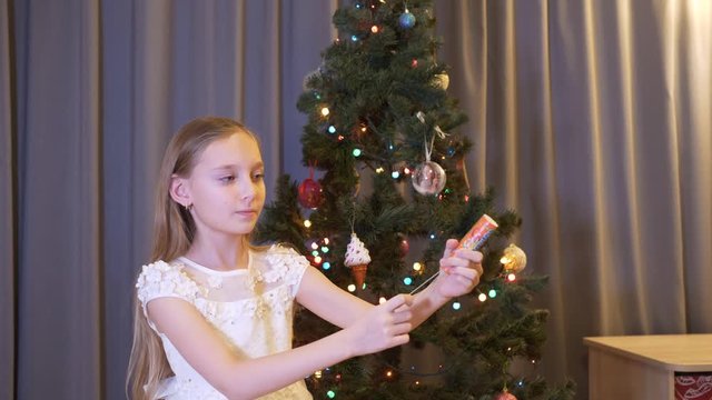 Scared little girl banging with cracker indoors at christmastime. Adorable frightened, little girl holding festive cracker and standing near christmas tree at home