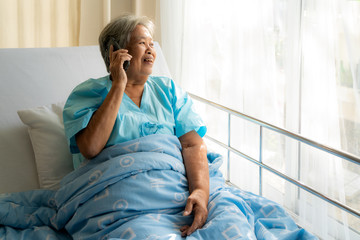 Asian senior elderly using chatting to family on her mobile phone and smiling, with delight as she listens to the conversation while she illness in hospital.