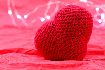 Valentine's day background. Background of crumpled red paper. Red knitted heart on a background of garland lights. Close-up, horizontal, side view, free space for text.