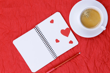 Background of Valentine's Day. On a red background in the center is a white notebook with hearts, a red pen. A cup of coffee in a white cup at the top right. Background of red crumpled paper. Flat lay