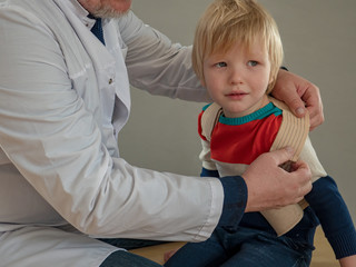 elderly doctor with gray hair and glasses in white coat and cap examines cute little blond boy in bright clothes with stethoscope. The doctor examines throat and listens to lungs. child trusts doctor