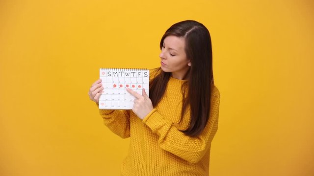 Young woman in bright sweater shirt hold in hand female periods calendar for checking menstruation days isolated over yellow background in studio. Medical healthcare gynecological lifestyle concept.
