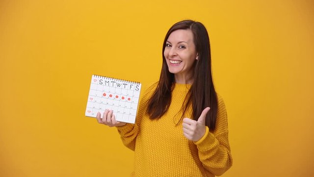 Young woman in sweater hold in hand female periods calendar for checking menstruation days show thumb up isolated over yellow background in studio. Medical healthcare gynecological lifestyle concept.