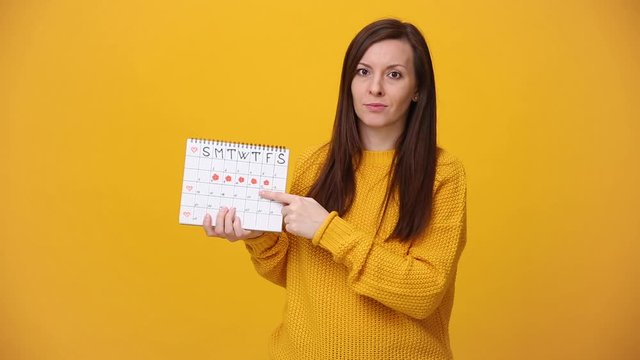 Young confused upset woman in sweater hold in hand female periods calendar for checking menstruation days isolated over yellow background in studio. Medical healthcare gynecological lifestyle concept.