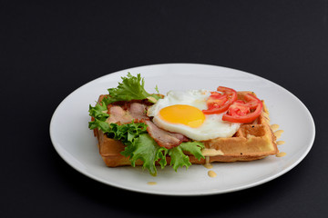 Classic eggs and bacon meat on waffles breakfast with tomatoes and salad served on a white plate on black background.