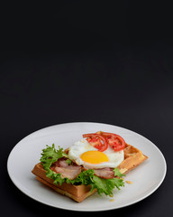 Classic eggs and bacon meat on waffles breakfast with tomatoes and salad served on a white plate on black background.