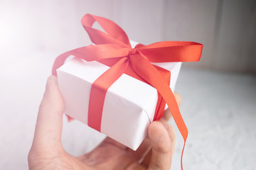 Hand holds gift box for Valentine's Day. View of present box package decorated into the white paper and red bow-knot. Flat lay on white background. ..