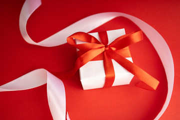 Top view of present box package decorated into the white paper and red bow-knot with white satin ribbons. Flat lay on red background. Concept of celebrate.