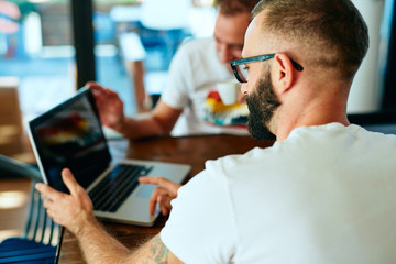 Attractive bearded man with glasses in a cafe working on laptop and chatting with a friend	