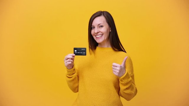 Smiling beautiful brunette young woman in bright sweater shirt posing isolated over yellow orange background in studio. People lifestyle concept. Hold in hand credit bank card, pointing with finger.