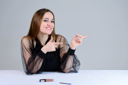 Woman is smiling beautifully at the camera, shows emotions. Photo of a cute brunette with excellent makeup in dark clothes sitting at a white table on a white background.