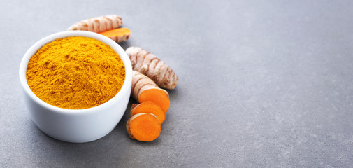 Turmeric root and bowl with turmeric powder on gray texture.