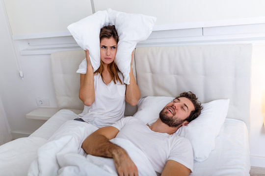 Young couple have problem with man's snoring. Heterosexual couple in bed, man sleeps and snoring with mouth open, while a tired woman irritated by snoring sitting on bed with a pillow on her head.