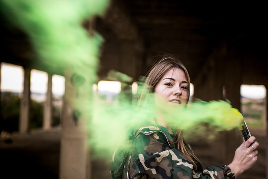Brown hair girl holds a smoke bomb that throws yellow smoke around. Young girl wears an urban style, military jacket and piercing.