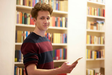 Teenager in library staying in front of bookshelf with smartphone in his hand