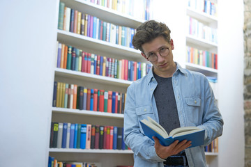 Serious young man with glasses, looking up from reading, next to a bookshelf