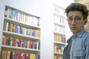 Young student with glasses, looking helpless and overcharged into the camera in library at...