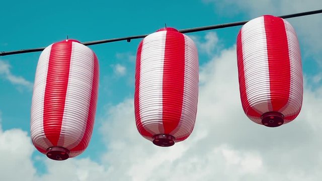 Oriental paper red-white lanterns Chochin hanging on white cloudy blue sky background. Japanese lanterns hanging on bright sky background. Pan right