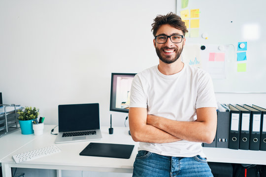 Portrait of confident graphic designer leaning on desk in office with arms crossed