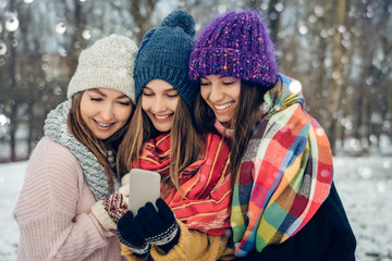 Three women friends outdoors in knitted hats using mobile phone on a snowy cold weather. Group of young female friends outdoors in winter park.