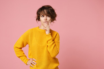 Pensive young brunette woman girl in yellow sweater posing isolated on pastel pink background studio portrait. People lifestyle concept. Mock up copy space. Put hand prop up on chin, looking aside.