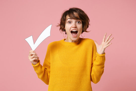 Excited young brunette woman girl in yellow sweater posing isolated on pastel pink wall background studio portrait. People lifestyle concept. Mock up copy space. Holding check mark, spreading hands.