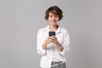 Smiling young business woman in white shirt posing isolated on grey background studio portrait. Achievement career wealth business concept. Mock up copy space. Using mobile phone, typing sms message.