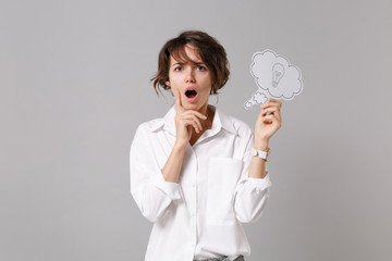 Shocked young business woman in white shirt posing isolated on grey background. Achievement career wealth business concept. Mock up copy space. Hold say cloud with lightbulb, put hand prop up on chin.