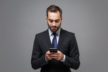 Handsome young business man in classic black suit shirt tie posing isolated on grey background. Achievement career wealth business concept. Mock up copy space. Using mobile phone typing sms message.