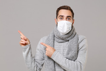Young man in gray sweater, scarf isolated on grey background. Healthy lifestyle, ill disease treatment, cold season concept. Mock up copy space. Wearing sterile face mask point index fingers aside up.